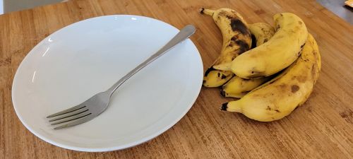 Ein leerer Teller steht auf einem Holztisch, darauf liegt eine Gabel, daneben liegen vier Bananen