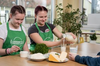 Zwei Teenager sitzen am Tisch, eine füllt ein Glas mit Milch, die andere rührt mit einem Löfel im Quark