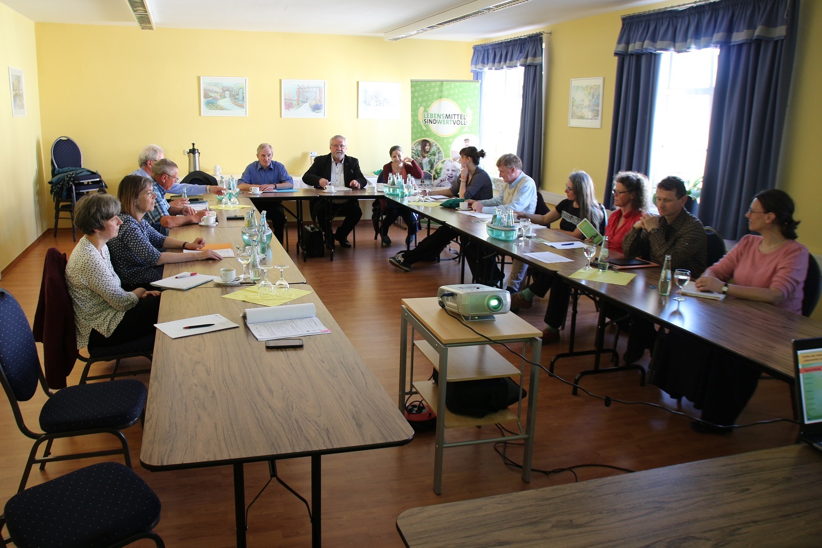 an U-förmig aufgestellten Tischen sitzen mehrere Personen und halten eine Konferenz